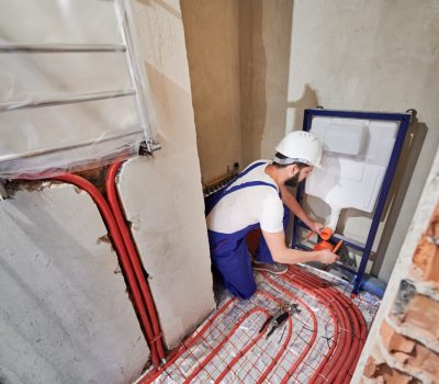 Wide angle photography of young plumber working in unfinished apartment, installing flush tank. Man wearing blue uniform and white helmet. Red pipes on the floor, new hanging radiator on the wall